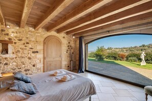 Ein Schlafzimmer mit Steinwand, Holzbalken, einem Bett mit Handtüchern und Blick auf eine Straße und Grünflächen.