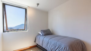 Una piccola camera con un letto singolo, una finestra, pavimento in legno e vista su una montagna in lontananza.