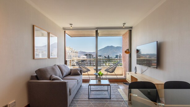 Un soggiorno moderno con divano, tavolino e televisione. È presente un balcone con vista su edifici e montagne.