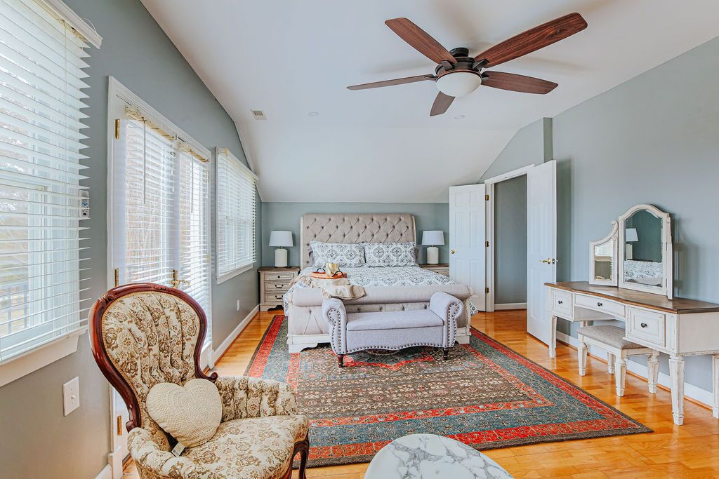 A bedroom with a bed, a chair, a desk, a ceiling fan, and a large window with blinds.