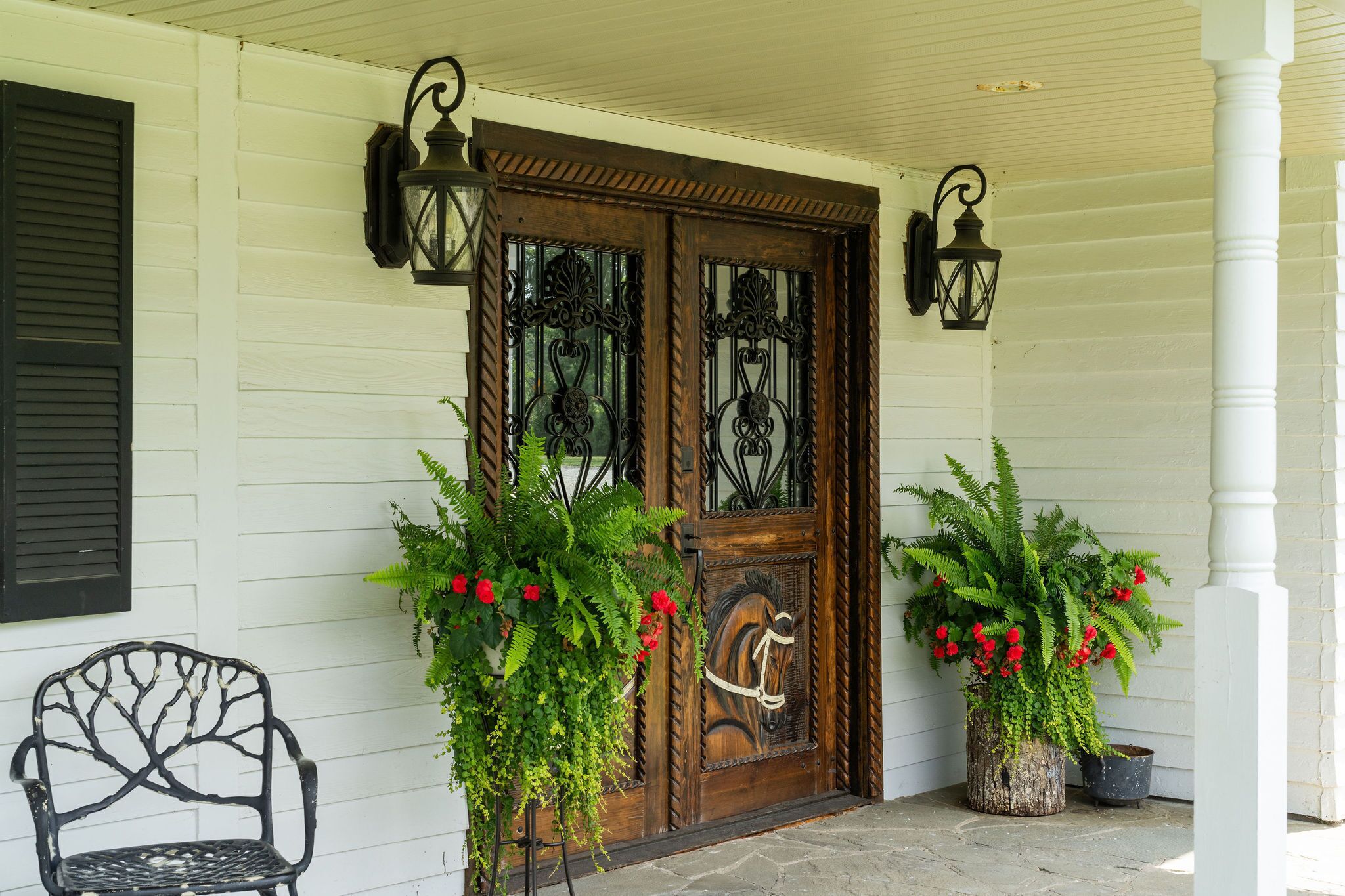 A wooden front door with decorative glass panels, flanked by two potted plants with red flowers, and a metal chair to the left.