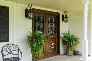 Uma porta de madeira com painéis de vidro decorativos, acompanhada por dois vasos com plantas e flores vermelhas, e uma cadeira de metal à esquerda.