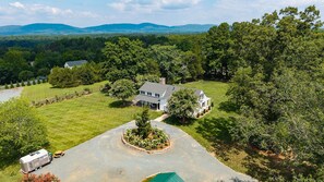 A two-story house with a large lawn, surrounded by trees and hills in the background.
