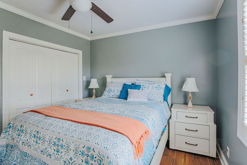 Une chambre à coucher comprenant un lit, deux tables de chevet, un ventilateur de plafond et une fenêtre avec des stores.