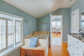 Une salle de bain avec une baignoire, un lavabo et un meuble-lavabo. Il y a une fenêtre avec des stores, une serviette accrochée à la baignoire et une chaise au fond de la pièce.