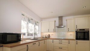 A modern kitchen with white cabinets, a microwave, a stove, and a window with a plant curtain. - Bluebell Cottage (Maidstone)