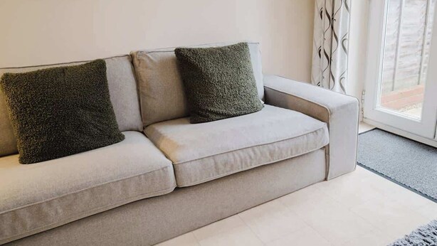 A light-colored sofa with two textured pillows, a plain wall, and a window with patterned curtains. - Spacious 4 Bed House With 3 Bathrooms & Parking (Bedford)