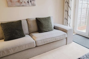 A light-colored sofa with two textured pillows, a plain wall, and a window with patterned curtains. - Spacious 4 Bed House With 3 Bathrooms & Parking (Bedford)