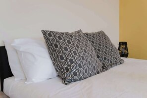 Una cama con un almohadón estampado en blanco y gris, una almohada blanca lisa y un objeto negro al lado.