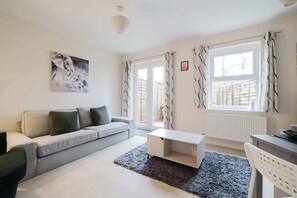 A modern living room with a grey sofa, a white coffee table, and a grey rug. There is a painting on the wall and a window with curtains. - Spacious 4 Bed House With 3 Bathrooms & Parking (Bedford)