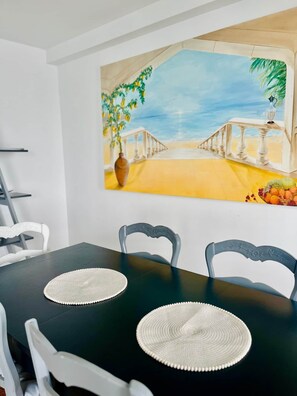 Un coin repas avec une table noire, des chaises blanches et un tableau représentant une scène de plage.