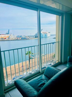 Un balcón con vista al puerto, que incluye un barco con la inscripción 'GNV' y una playa con palmeras.