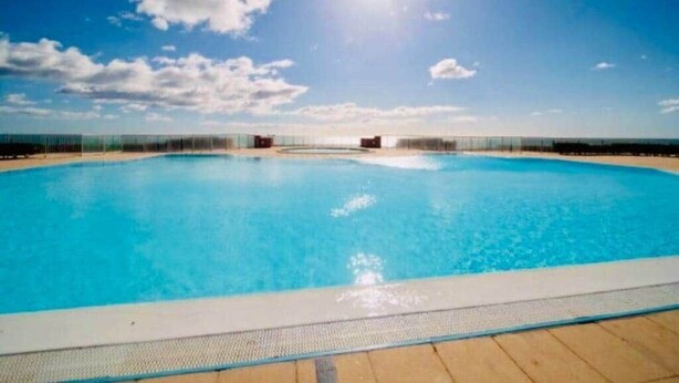 A clear blue swimming pool with a wooden deck, surrounded by a fence, under a sunny sky.