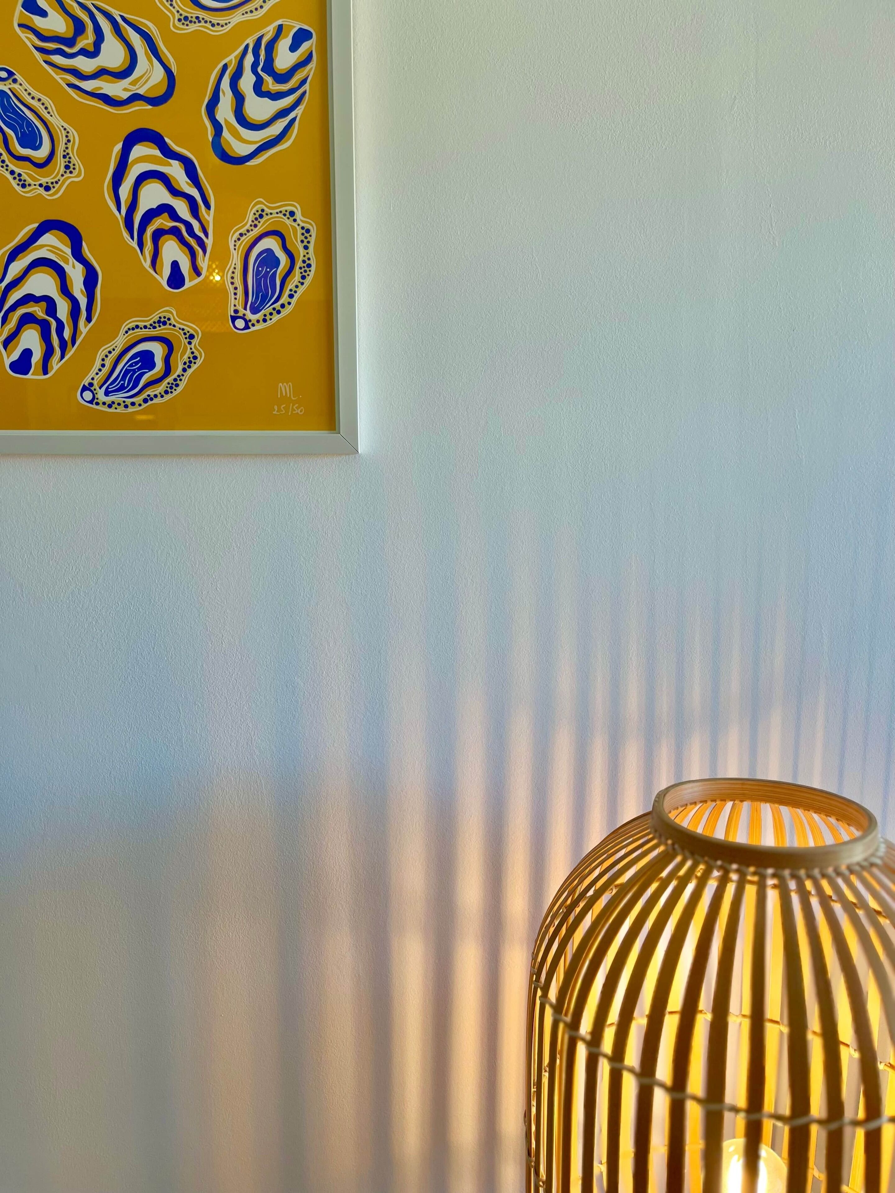 Ein gerahmtes Kunstwerk mit abstrakten blauen und weißen Mustern auf gelbem Grund, eine strukturierte weiße Wand und eine Lampe mit einer gerippten, lichtdurchlässigen Schirm.
