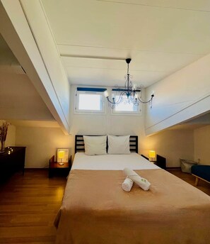 Una habitación de hotel con una cama, un candelabro, un ventanal pequeño y una mesita de noche con lámpara.