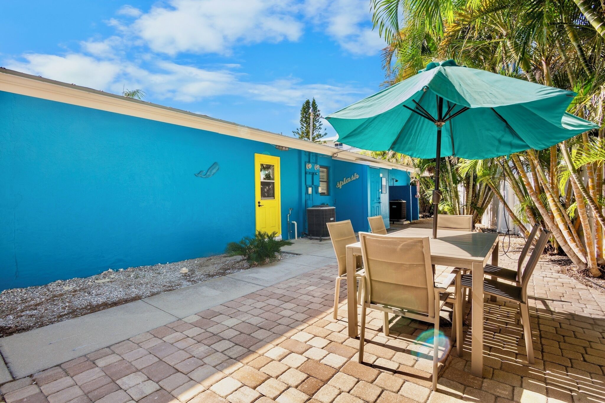 A blue building with a yellow door, outdoor seating with a green umbrella, and palm trees.