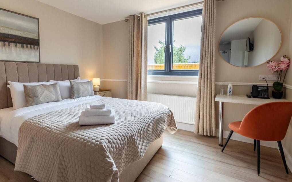 Habitación de hotel con cama, escritorio, silla, ventana con cortinas y un espejo redondo en la pared.