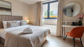 Habitación de hotel con cama, escritorio, silla, ventana con cortinas y un espejo redondo en la pared.
