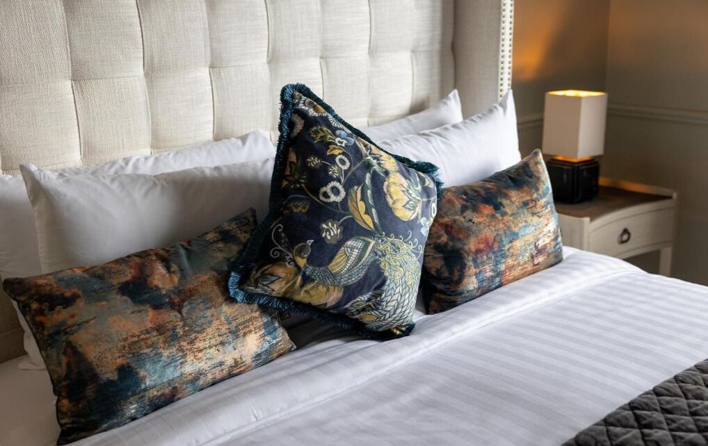 Una cama con ropa de cama blanca y almohadas decorativas, una mesita de noche con lámpara y un cabecero capitonado.