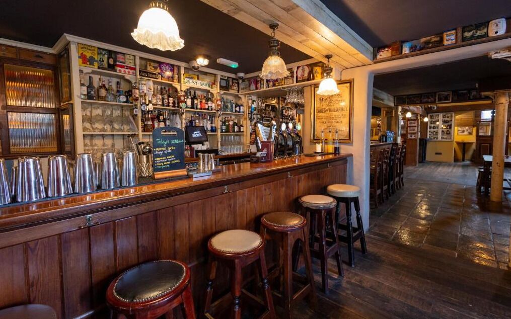 Una zona de bar con mostradores de madera, taburetes y una variedad de botellas y vasos.