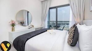 A modern hotel room with a large bed, a round mirror, a view of the city through large windows, and a balcony with a table and chairs. - Vogue | Sobha Hartland close to Downtown Dubai Mall | Burj Khalifa (Dubai)