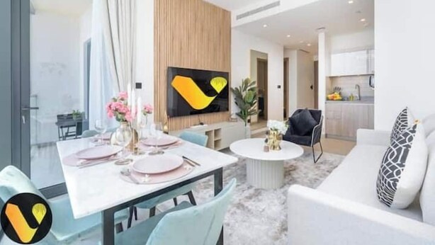 A modern living room with a dining area, a kitchen, and a large TV mounted on the wall. - Vogue | Sobha Hartland close to Downtown Dubai Mall | Burj Khalifa (Dubai)