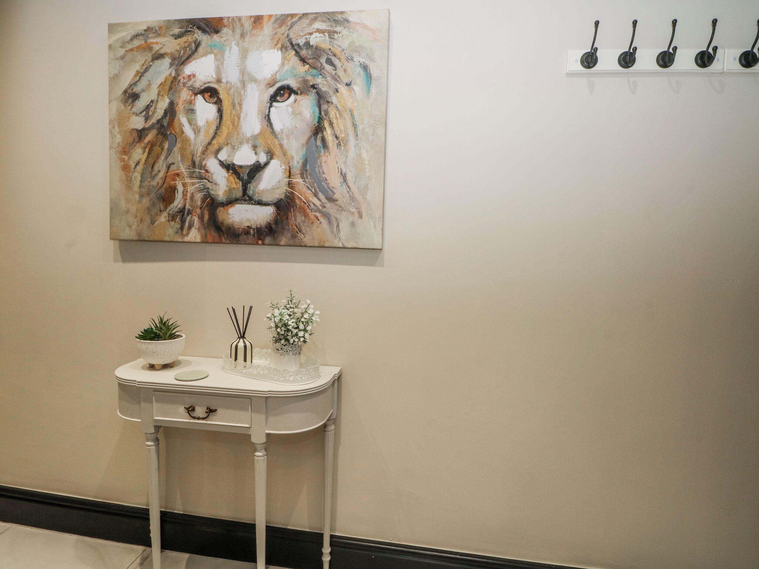 A bathroom with a painting of a lion's face, a small table with a plant and a vase, and a row of hooks on the wall.