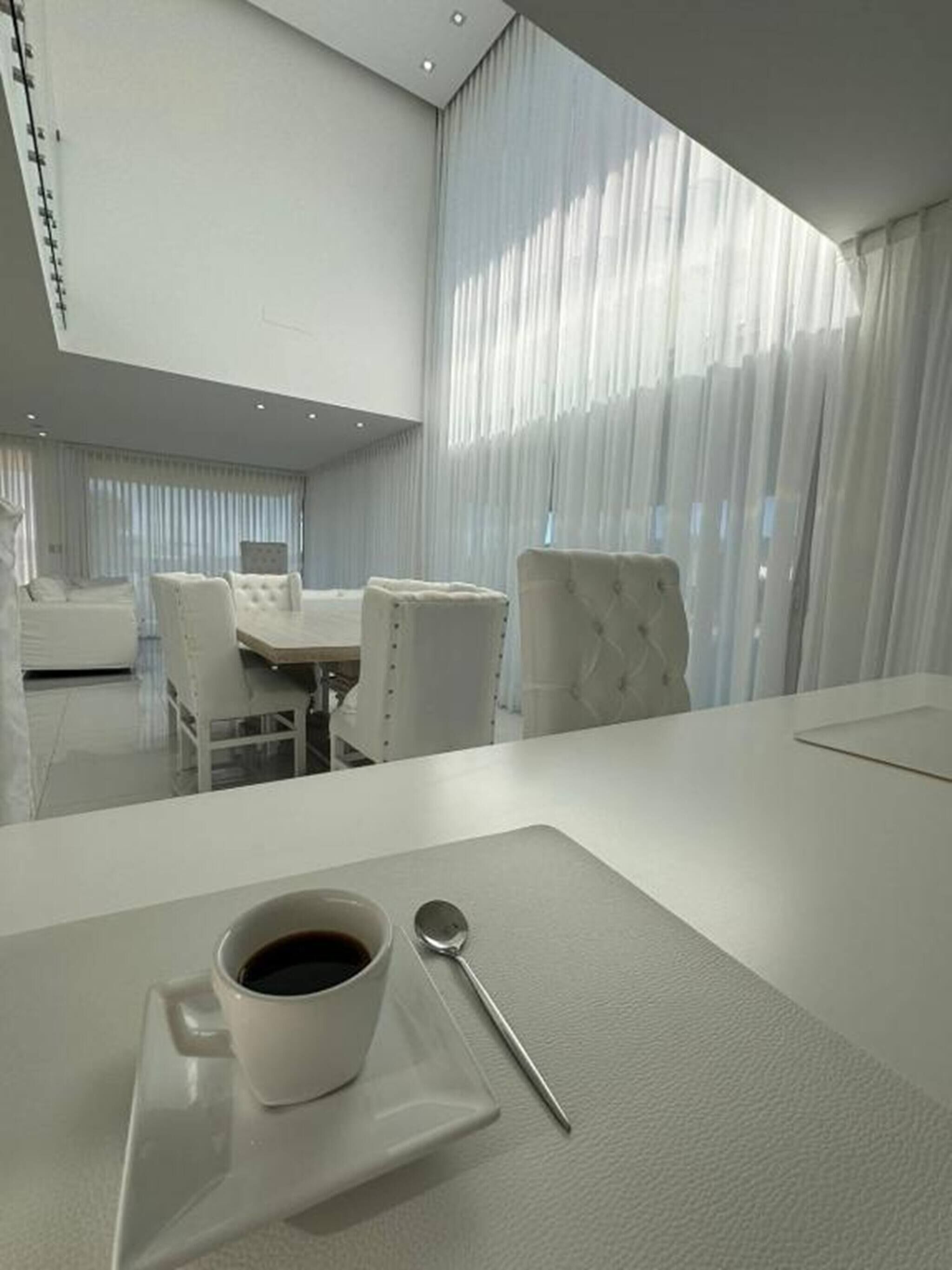 A modern hotel room with a dining area, a coffee table with a cup and spoon, and sheer curtains allowing natural light.