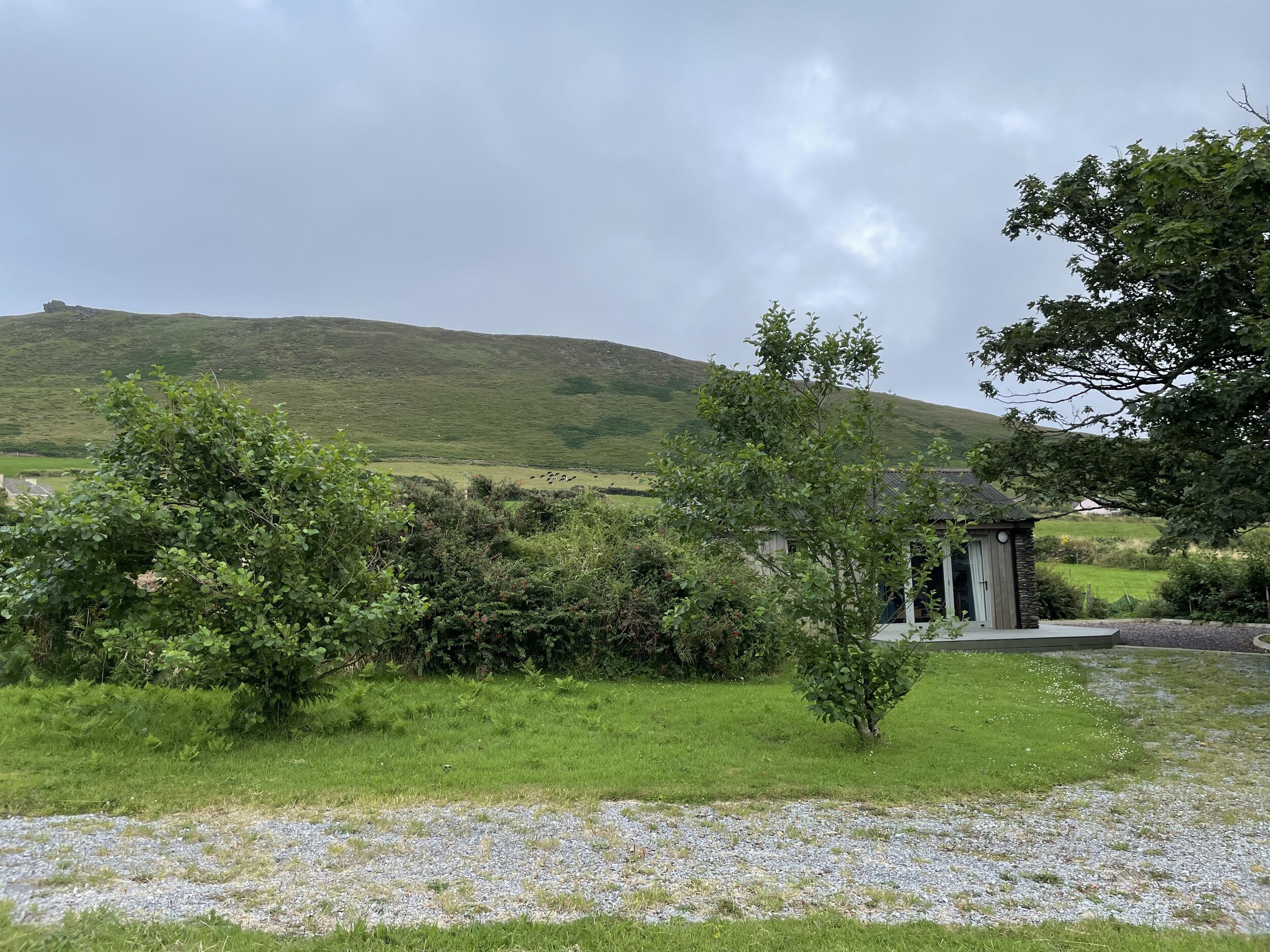 A rural landscape with a small stone building, surrounded by greenery and a hill in the background.