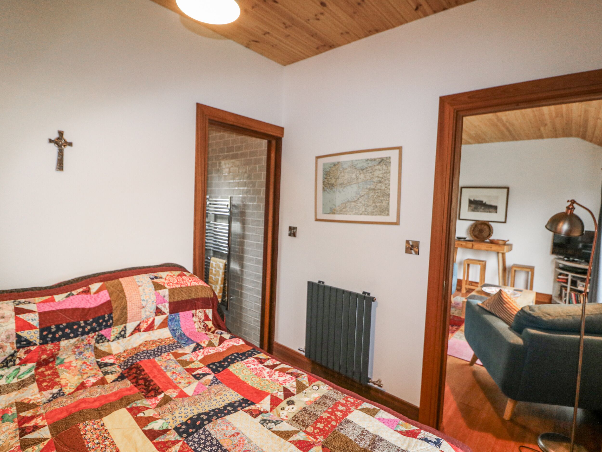 Un dormitorio con una cama grande, un radiador, techo de madera, una cruz en la pared y una puerta que da a otra habitación.