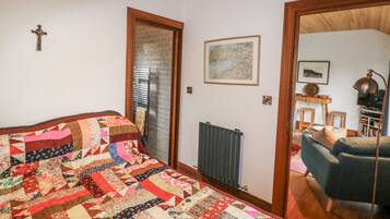 Un dormitorio con una cama grande, un radiador, techo de madera, una cruz en la pared y una puerta que da a otra habitación.