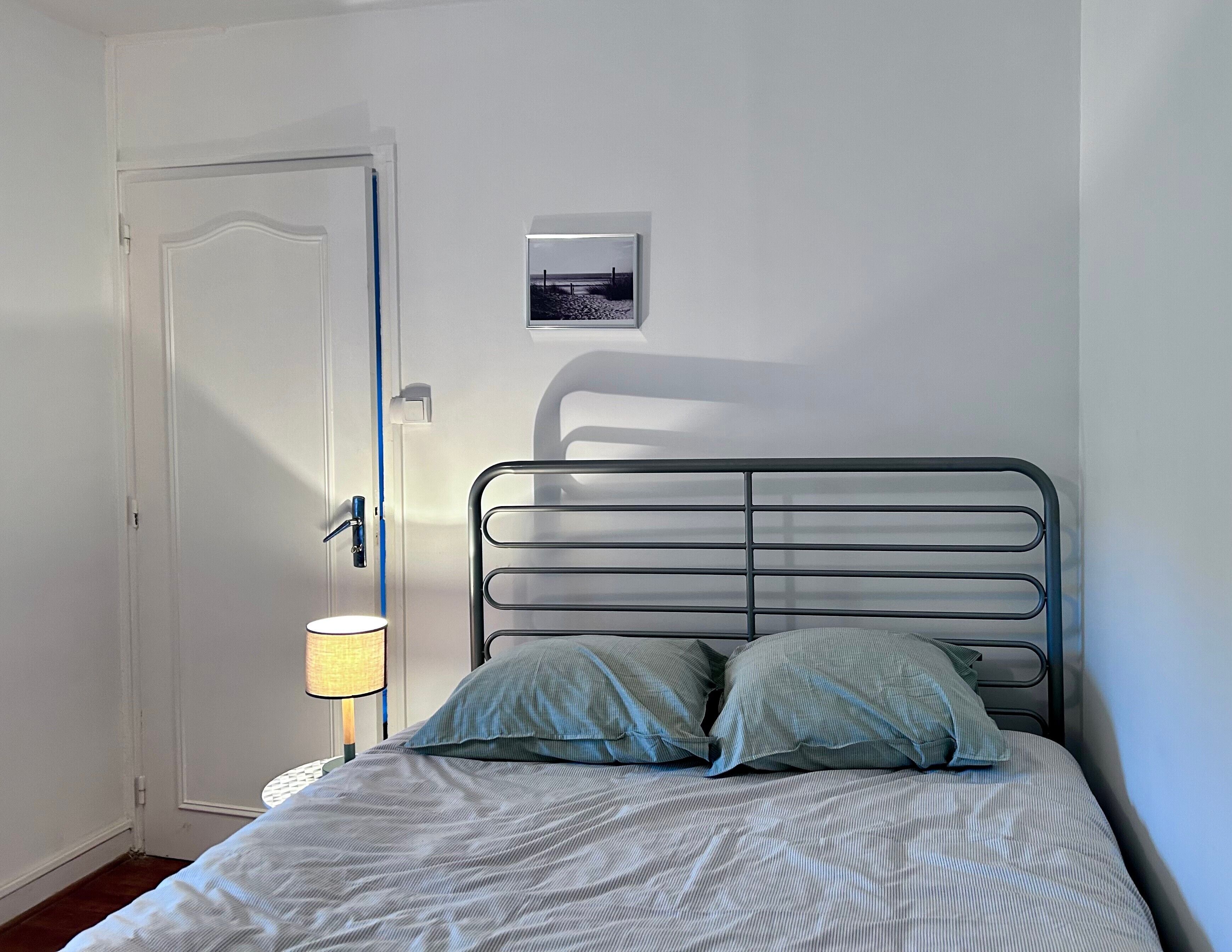 A bedroom with a bed, a door, a wall-mounted picture, and a bedside lamp.