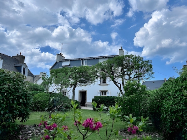 A white building with a dark roof, surrounded by lush greenery and flowering plants. - Treboul – Family Home Close to Beaches and Shops (Douarnenez)