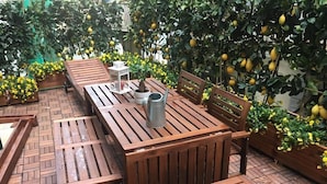 Ett utomhusbord och stolar i trä, placerade på en innergård med ett stengolv, omgivet av krukväxter och med citronträd i bakgrunden.