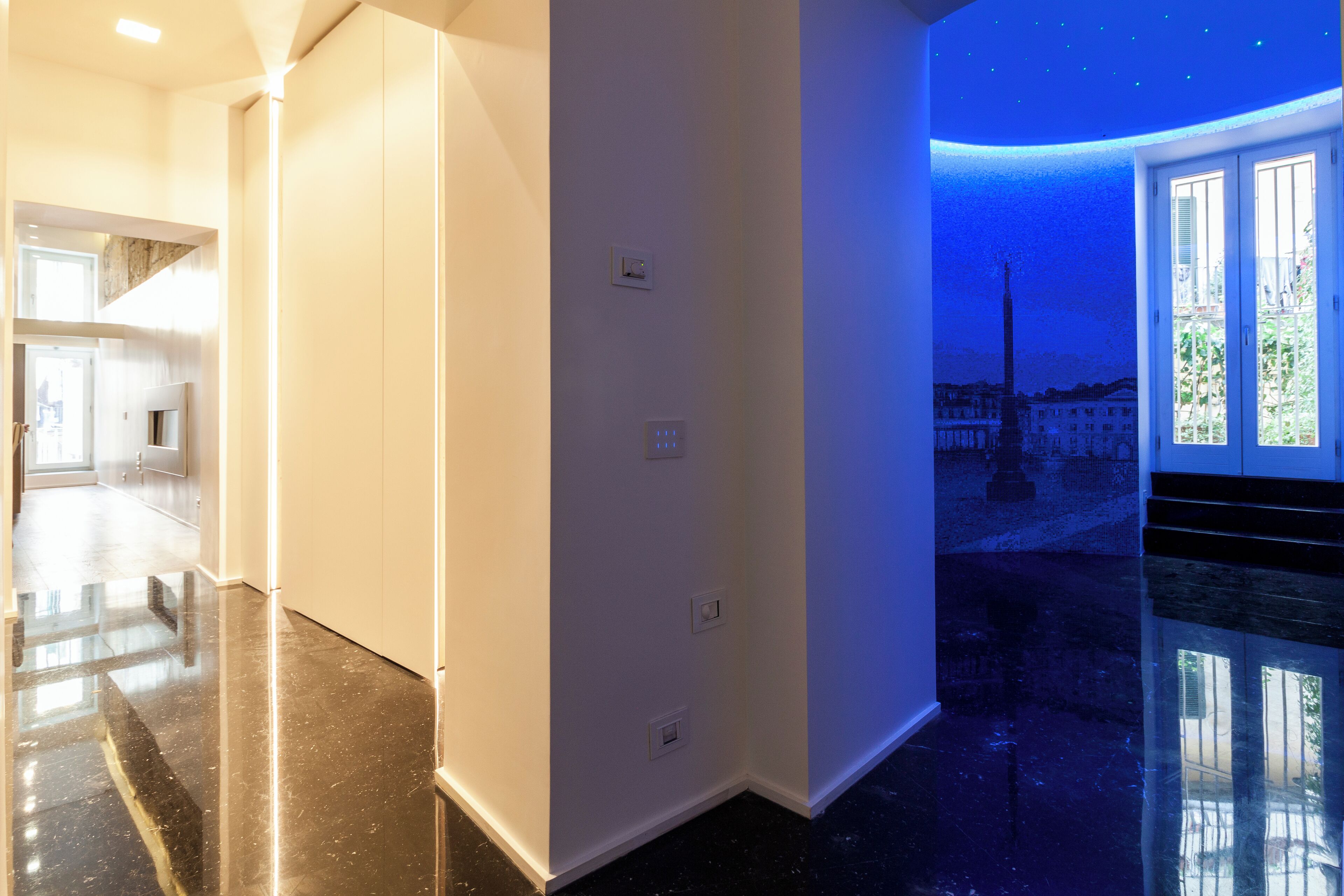 A modern hotel interior with a spacious hallway, a large white wall with electrical outlets, and a view of a cityscape through a glass door.