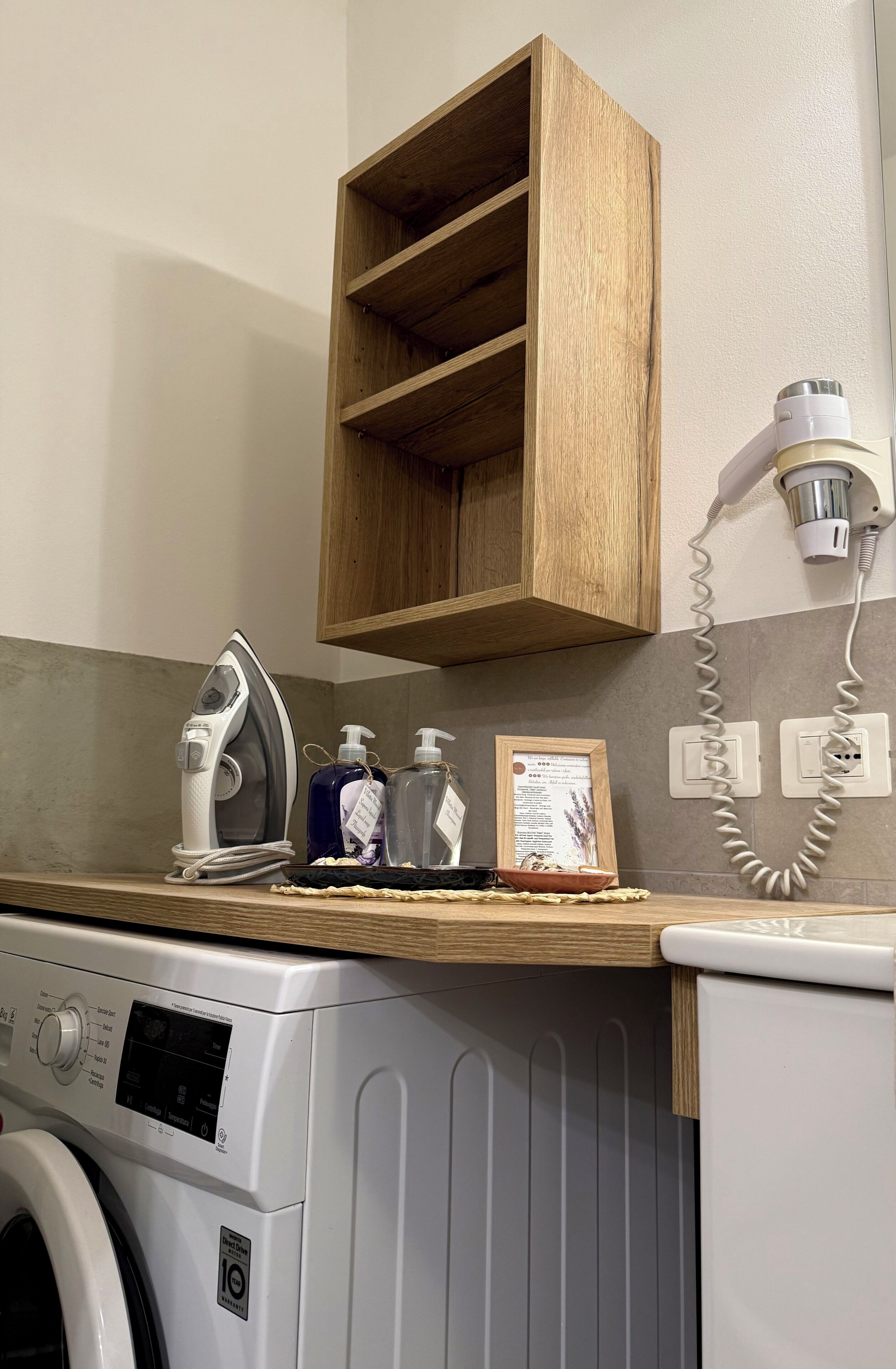 A laundry area with a washing machine, an iron, and a wooden shelf above.