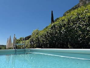 A clear swimming pool with a metal ladder, surrounded by lush greenery and a clear blue sky. - Villetta Moscal- Buon Gusto (Affi)