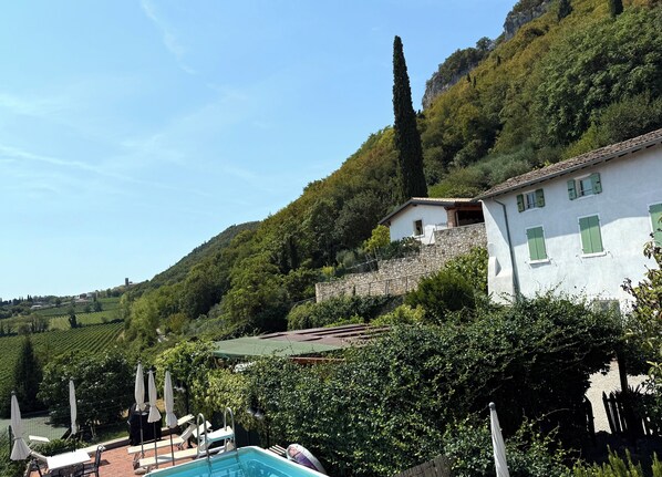 Zona de piscina con sillas de descanso y sombrillas, un muro de piedra y un edificio con fachada de piedra y contraventanas verdes.