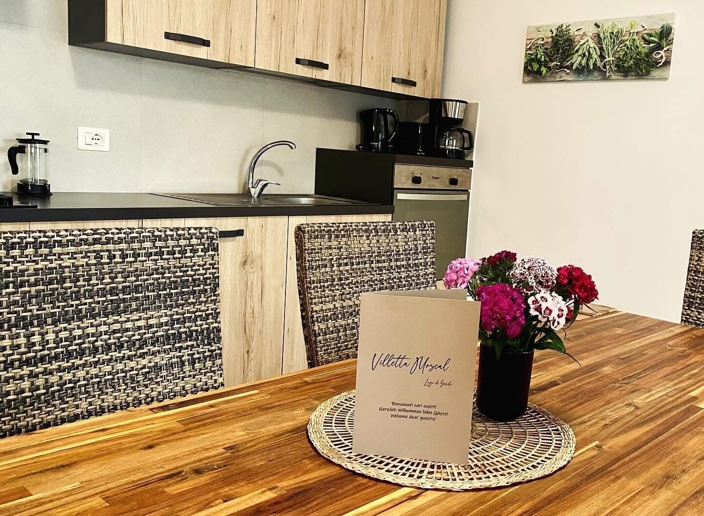 A modern kitchen with a sink, stove, and coffee maker. There is a wooden dining table with a placemat and a vase of flowers. A welcome sign is on the table.