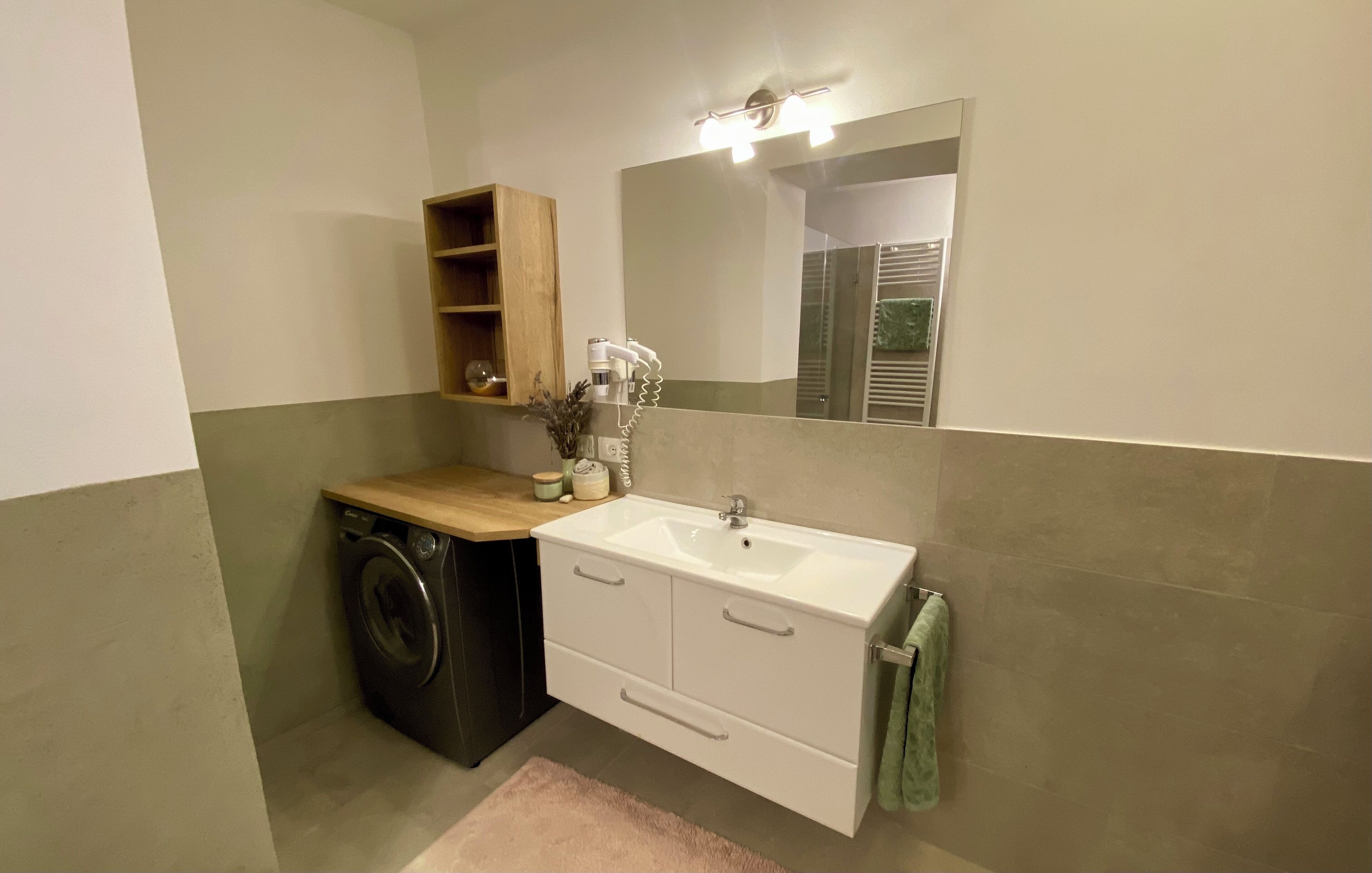 A compact bathroom with a washing machine, a sink, a mirror, and a wooden shelf.