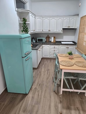 Une cuisine compacte avec des meubles blancs, un réfrigérateur de couleur turquoise, un four à micro-ondes et une table à manger agrémentée de sets de table en fibres tressées.