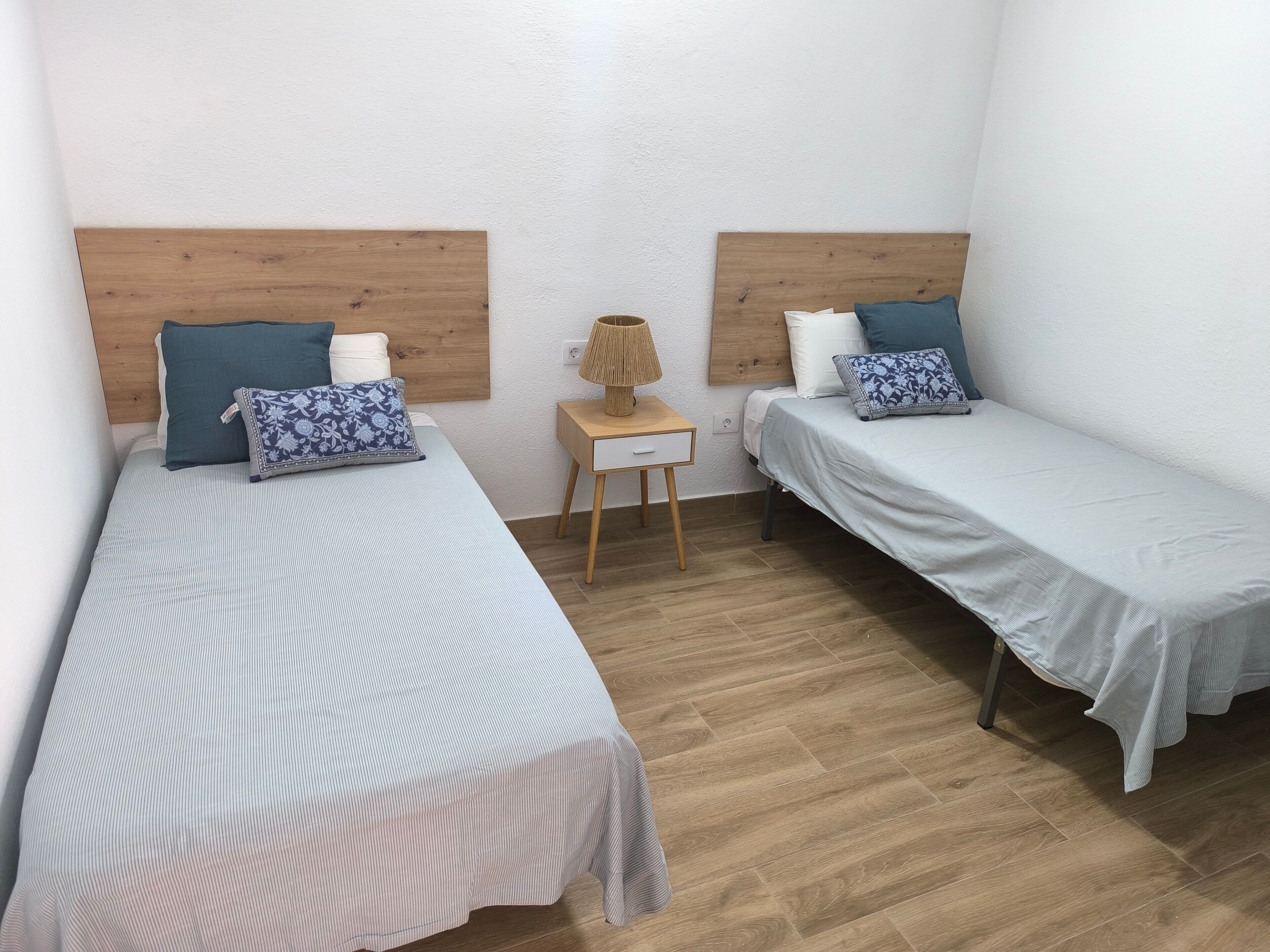 A room with two single beds, a small wooden nightstand with a lamp, and a white wall.