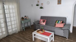 Una sala de estar con un sofá gris, una mesa de centro blanca, una mesita auxiliar y un ventanal grande con persianas.