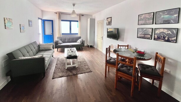 Un salon comprenant un canapé, une table basse, une table à manger avec des chaises, un téléviseur fixé au mur et une fenêtre avec des rideaux.