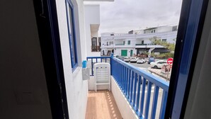 Un balcon étroit avec une balustrade bleue, une table blanche et une vue sur une rue bordée de voitures garées et d’immeubles.