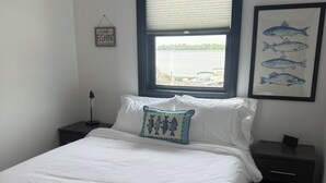 A bedroom with a bed, a nightstand, a window with blinds, and a framed fish picture on the wall. - Relaxing Lakeside Cabin Retreat on Lake Champlain (Rouses Point)