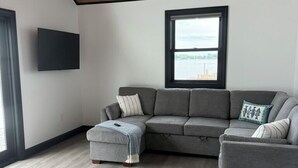 A modern living room with a sectional sofa, ottoman, and a large window. - Relaxing Lakeside Cabin Retreat on Lake Champlain (Rouses Point)