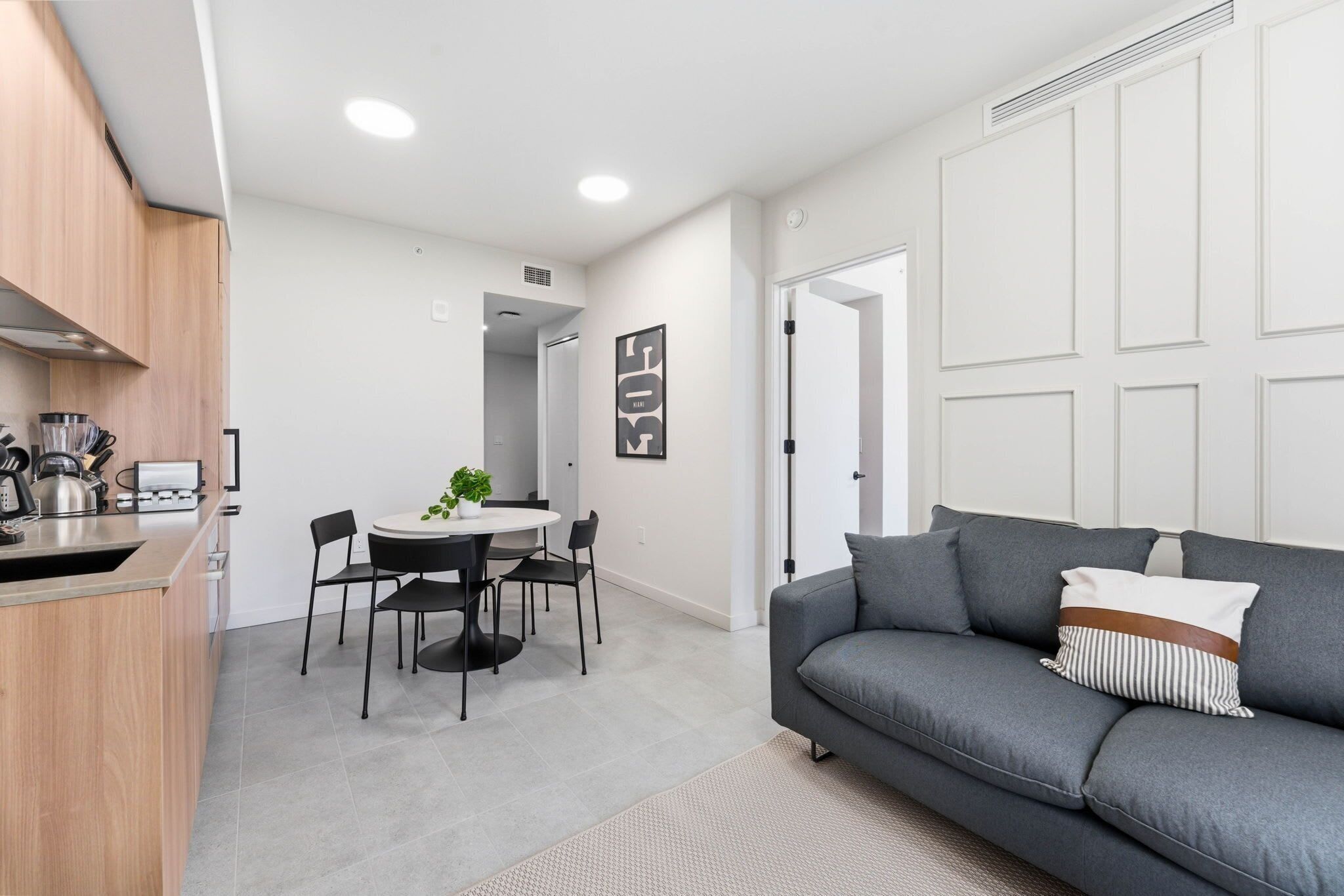 Una sala de estar moderna con un sofá gris, una mesa de comedor con sillas, y una cocina con armarios de madera y un hervidor.
