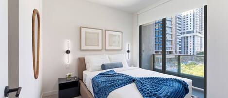 A modern bedroom with a large window, a bed with a blue blanket, a nightstand, and two wall-mounted lights.