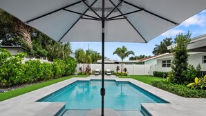 A rectangular swimming pool with a white umbrella overhead, surrounded by a well-maintained lawn and various plants. - Turfside Terrace, Heated Pool and Putt Putt (Jupiter)